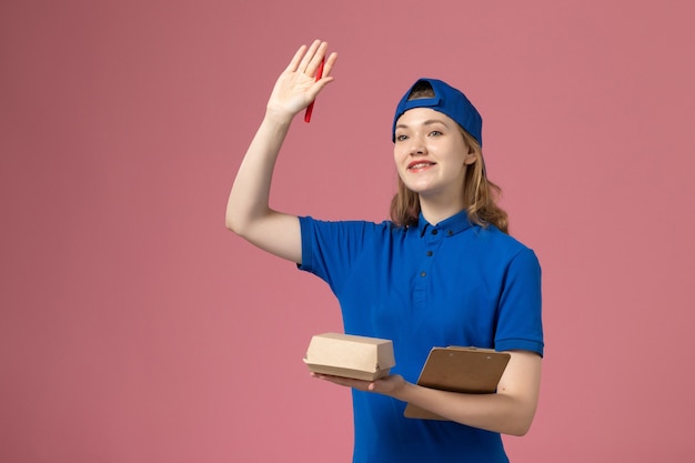 Женщина-курьер в синей форме и накидке, держащая небольшой блокнот для доставки еды и пишущий на розовой стене, работа сотрудника службы доставки