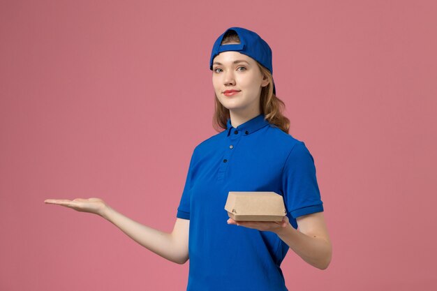 ピンクの背景の配達サービス会社の労働者の女の子の仕事に小さな配達食品パッケージを保持している青い制服と岬の正面図の女性の宅配便