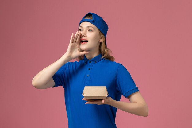 Front view female courier in blue uniform and cape holding little delivery food package on the pink background delivery uniform service company work girl job