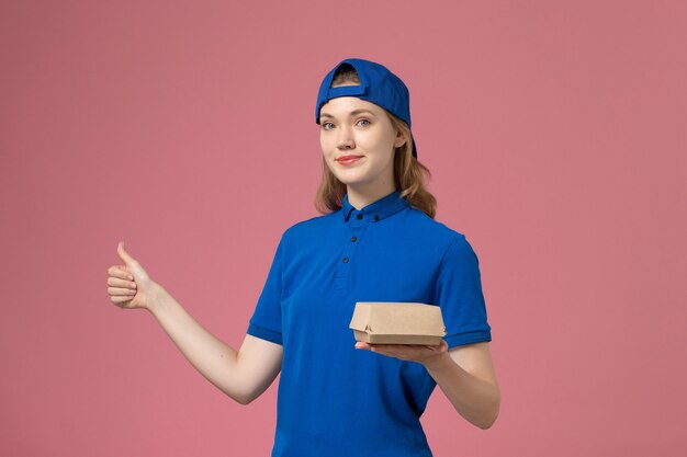 Front view female courier in blue uniform and cape holding little delivery food package on the pink background delivery uniform service company work job
