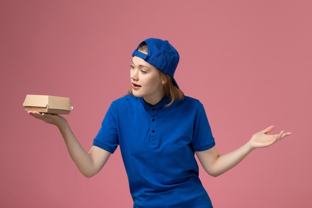 Corriere femminile di vista frontale in uniforme blu e mantello che tiene piccolo pacchetto di cibo di consegna su sfondo rosa lavoratore di società di servizio uniforme di consegna di lavoro