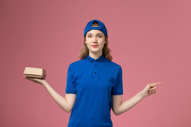 Free photo front view female courier in blue uniform and cape holding little delivery food package on pink background job delivery work uniform service company