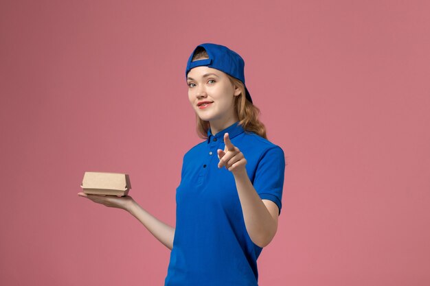 Front view female courier in blue uniform and cape holding little delivery food package on pink desk job delivery uniform service work company