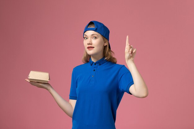 Front view female courier in blue uniform and cape holding little delivery food package on pink wall, delivery job uniform service company