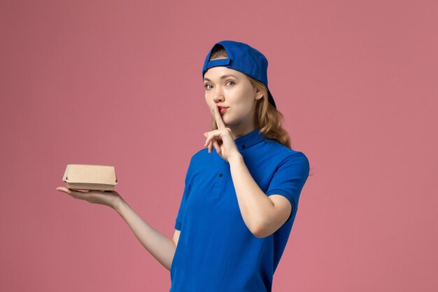 Front view female courier in blue uniform and cape holding little delivery food package on pink wall, delivery uniform service company worker girl
