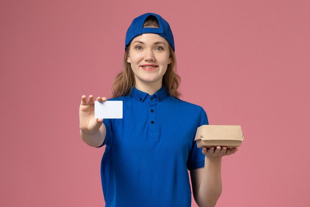 파란색 유니폼과 케이프 핑크 벽, 배달 작업 작업 서비스 직원에 카드와 함께 작은 배달 음식 패키지를 들고 전면보기 여성 택배