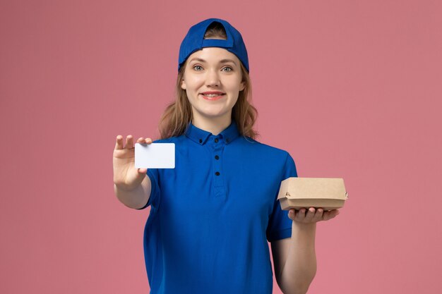 Front view female courier in blue uniform and cape holding little delivery food package with card on pink wall, delivery job work service employee