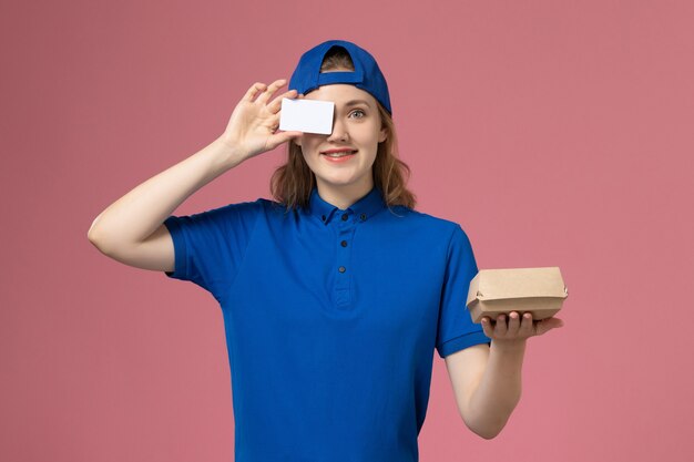 Женщина-курьер в синей униформе и накидке с карточкой на розовой стене, вид спереди, работа сотрудника службы доставки