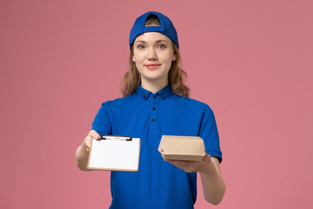 Front view female courier in blue uniform and cape holding little delivery food package with notepad on pink wall, job delivery service employee