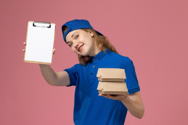 Front view female courier in blue uniform cape holding little delivery food packages and notepad on pink background job delivery service employee work