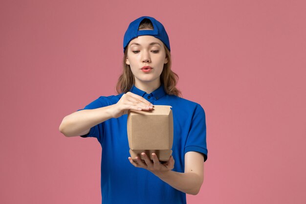 Front view female courier in blue uniform and cape holding little delivery food packages on the pink wall, delivery job uniform service employee worker