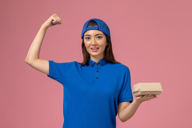 Курьер-женщина, вид спереди в синей форменной накидке, держит небольшую посылку для доставки и сгибается на розовой стене, служба доставки сотрудников