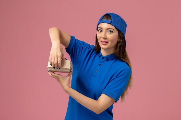 Front view female courier in blue uniform cape holding little delivery package opening it on pink wall, employee service delivery work job worker