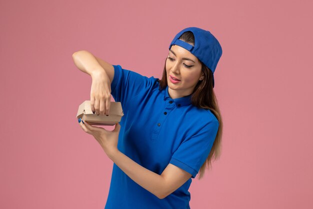 Front view female courier in blue uniform cape holding little delivery package on pink wall, employee service delivery company