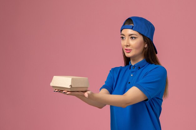 Front view female courier in blue uniform cape holding little delivery package on pink wall, employee service delivery work job worker girl