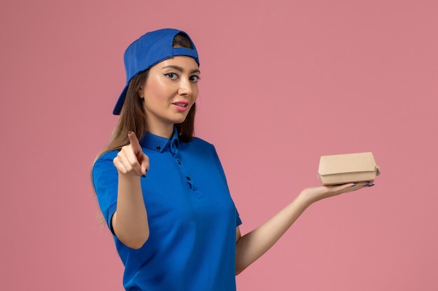 Front view female courier in blue uniform cape holding little delivery package on the pink wall, employee service delivery work job