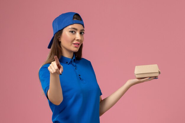Женщина-курьер в синей униформе, держащая небольшую посылку на розовой стене, вид спереди, работа по доставке сотрудников