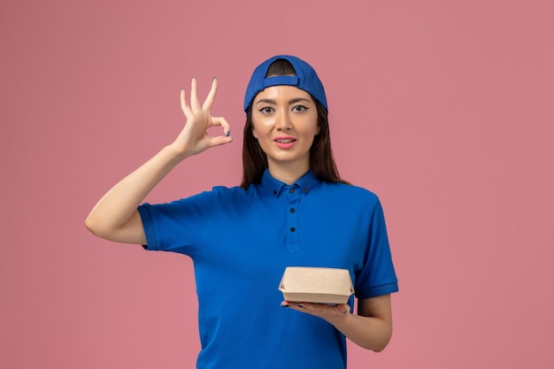 ピンクの壁に小さな配達パッケージを保持している青い制服の岬の正面図の女性の宅配便、従業員サービスの配達作業