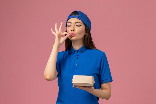 Front view female courier in blue uniform cape holding little delivery package on pink wall, employee service delivery