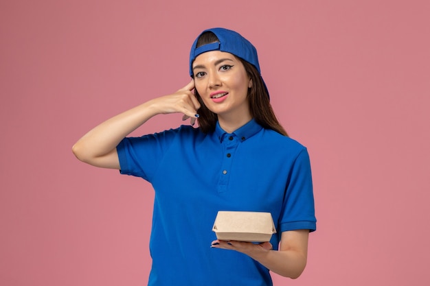 Front view female courier in blue uniform cape holding little delivery package on pink wall, employee service work job worker girl company