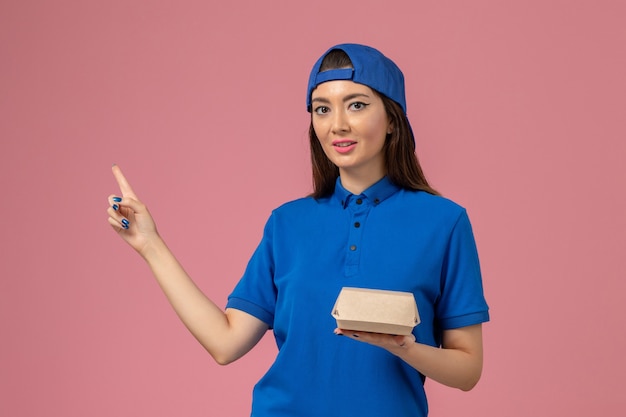 ピンクの壁に小さな配達パッケージを保持している青い制服の岬の正面図の女性の宅配便、サービス配達仕事の労働者の女の子の会社