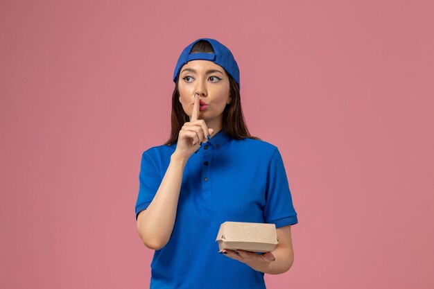 Женщина-курьер в синей форме, держащая небольшую посылку с доставкой и показывающая знак тишины на розовой стене, работа службы доставки сотрудников