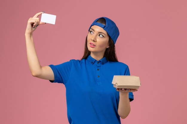 Front view female courier in blue uniform cape holding little delivery package with plastic card on light pink wall, employee service delivery work