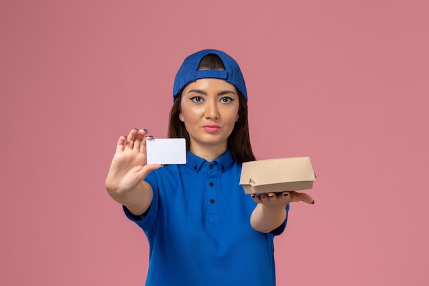 淡いピンクの壁にプラスチックカードと小さな配達パッケージを保持している青い制服の岬の正面図の女性の宅配便、従業員サービス配達労働者