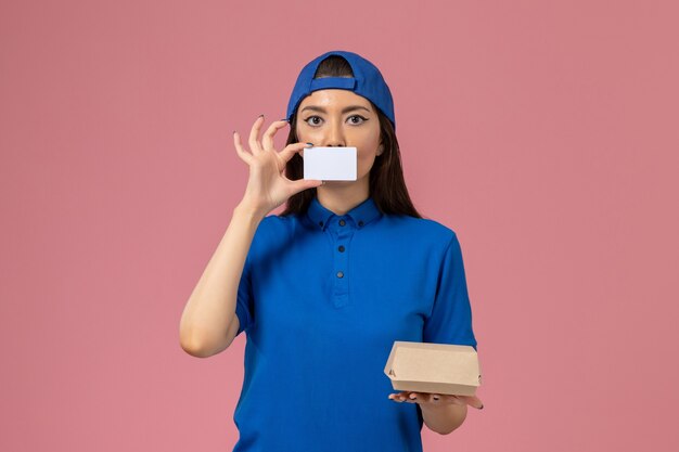 Front view female courier in blue uniform cape holding little delivery package with plastic card on light pink wall, employee work job service delivery