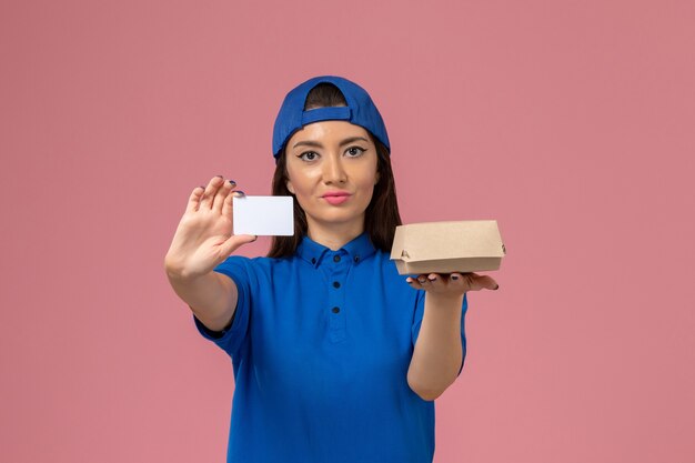 Front view female courier in blue uniform cape holding little delivery package with plastic card on light pink wall, work job employee service delivery