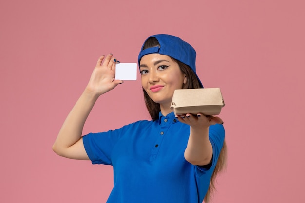 Front view female courier in blue uniform cape holding little delivery package with plastic card smiling on light pink wall, employee service delivery