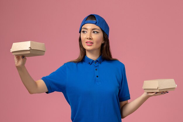 Front view female courier in blue uniform cape holding little delivery packages on pink wall, employee service company delivery