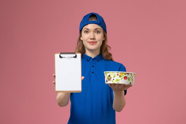 Front view female courier in blue uniform cape holding notepad and delivery bowl on pink wall, service delivery employee