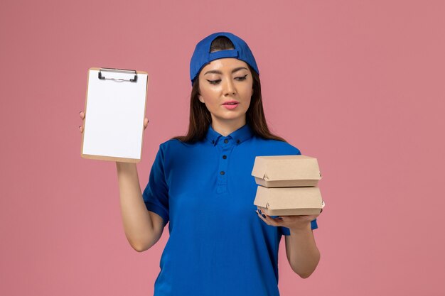 Женщина-курьер в синей форме, держащая блокнот и маленькие посылки на светло-розовой стене, работник службы доставки