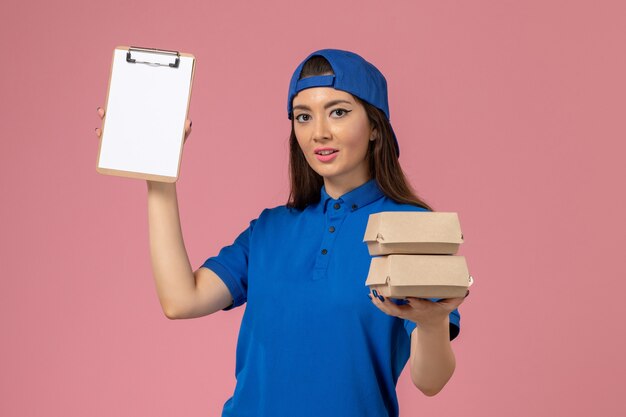 Front view female courier in blue uniform cape holding notepad and little delivery packages on light pink wall, service employee delivery