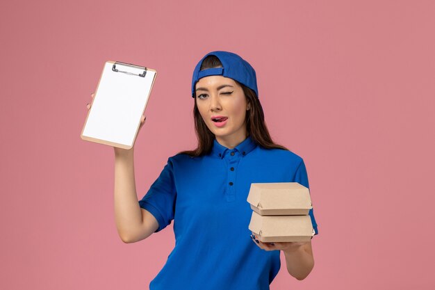 Front view female courier in blue uniform cape holding notepad and little delivery packages winking on light pink wall, service employee delivery