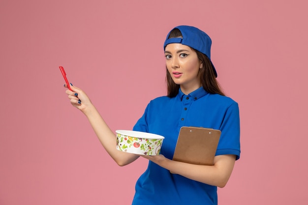 Женщина-курьер в синей форменной накидке, вид спереди, держит блокнот и ручку с миской на светло-розовой стене, служба доставки работает