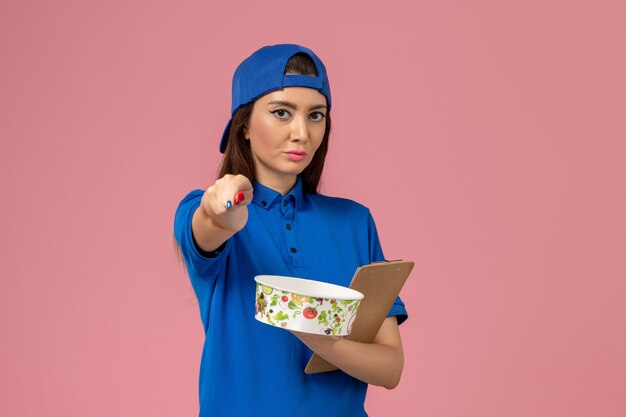 Front view female courier in blue uniform cape holding notepad and pen with delivery bowl on light-pink wall, service employee delivery worker