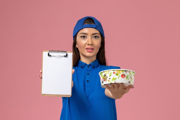 Front view female courier in blue uniform cape holding notepad with delivery bowl on light-pink wall, girl service employee delivery