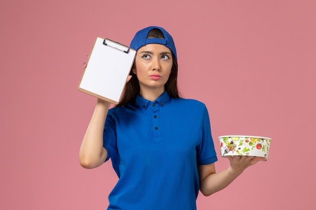 Front view female courier in blue uniform cape holding notepad with delivery bowl thinking on light-pink wall, service employee delivering job