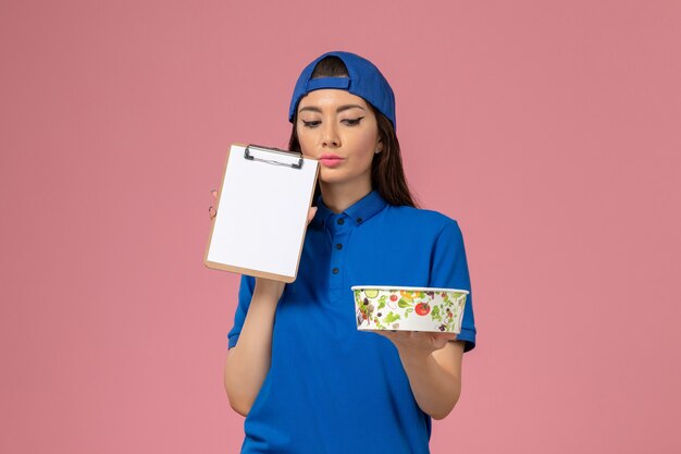 Front view female courier in blue uniform cape holding notepad with delivery bowl thinking on light-pink wall, service employee delivering