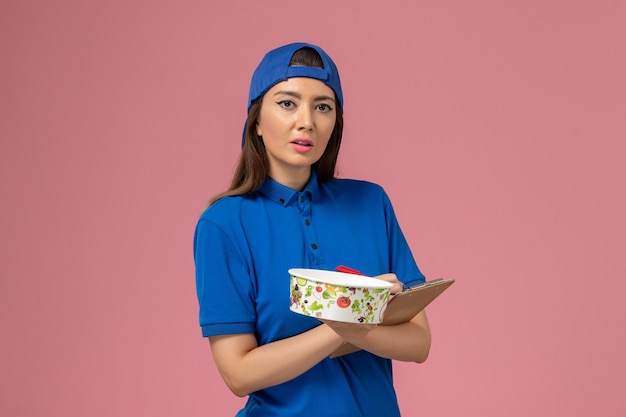 Front view female courier in blue uniform cape holding notepad with delivery bowl writing notes on light-pink wall, service job employee delivery