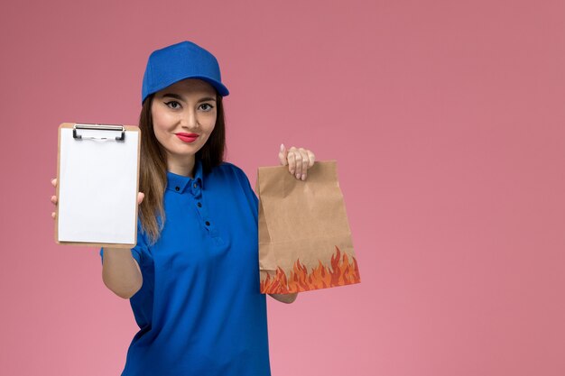 Front view female courier in blue uniform and cape holding notepad with paper food package on the pink desk