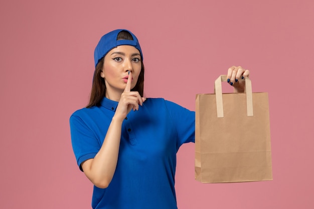 Front view female courier in blue uniform cape holding paper delivery package asking to be quiet on the light-pink wall, service employee delivering