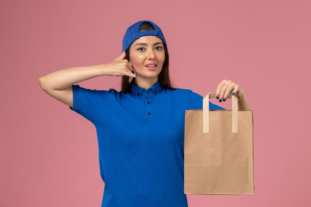 Front view female courier in blue uniform cape holding paper delivery package on the light-pink wall, service employee delivering job