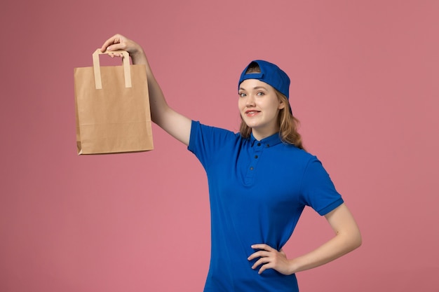 Front view female courier in blue uniform cape holding paper delivery package on pink wall, service delivery employee job
