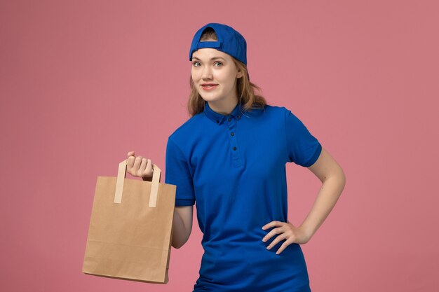 Front view female courier in blue uniform cape holding paper delivery package on pink wall, service delivery employee