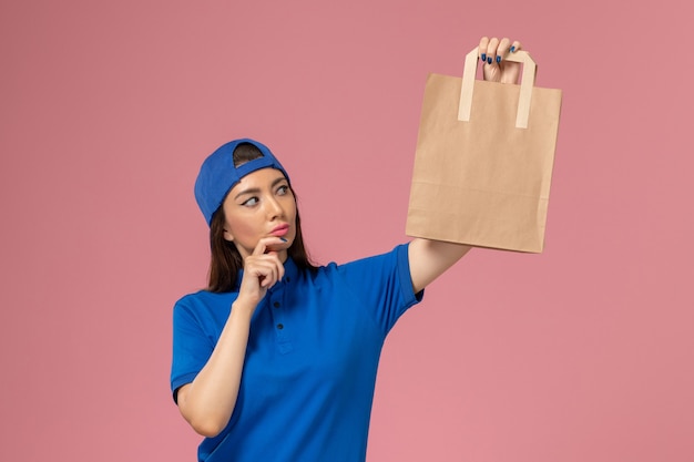 Front view female courier in blue uniform cape holding paper delivery package thinking on light-pink wall, service employee delivering