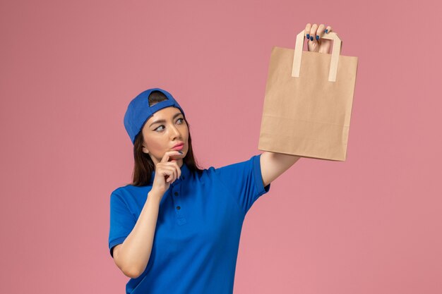 淡いピンクの壁を考えて紙の配達パッケージを保持している青い制服の岬の正面図の女性の宅配便、サービス従業員が配達