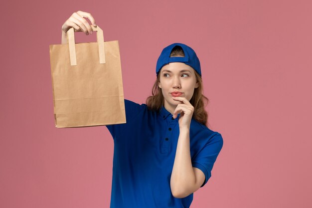 Женщина-курьер в синей форме, держащая пакет доставки бумаги, думает о розовой стене, сотрудник службы доставки